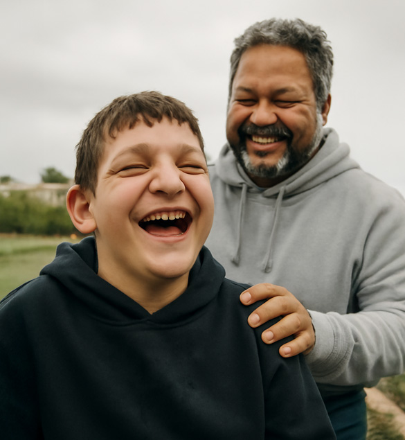 Una imagen de dos personas compartiendo un momento alegre al aire libre. Un joven sonr&iacute;e ampliamente mientras un hombre adulto, de pie detr&aacute;s de &eacute;l, lo observa con una expresi&oacute;n sonriente y cari&ntilde;osa. Ambos visten ropa c&oacute;moda y est&aacute;n rodeados de un ambiente natural con cielo nublado y &aacute;reas verdes al fondo.
