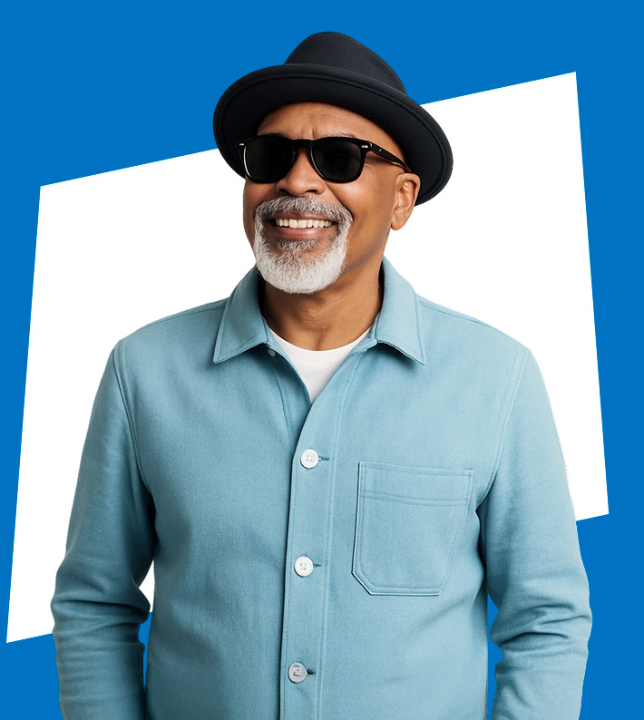 Hombre adulto mayor de piel morena, con barba y bigote blancos, usando sombrero negro y gafas de sol. Sonríe mientras viste una chaqueta celeste sobre una polera blanca.