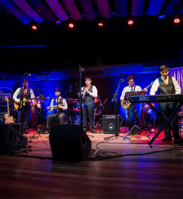 Banda en vivo sobre un escenario iluminado con luces azules y rojas. Seis músicos vestidos con camisas blancas y chalecos oscuros interpretan diferentes instrumentos: guitarra, charango, flauta, bajo, teclado y percusión. Al fondo se observa una batería y equipo de sonido. La escena transmite energía y ambiente festivo de concierto.